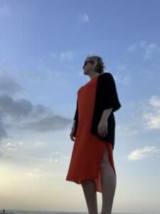 Katie na praia vestindo um longo vestido laranja com capa preta em cima para cobrir ombros e braços