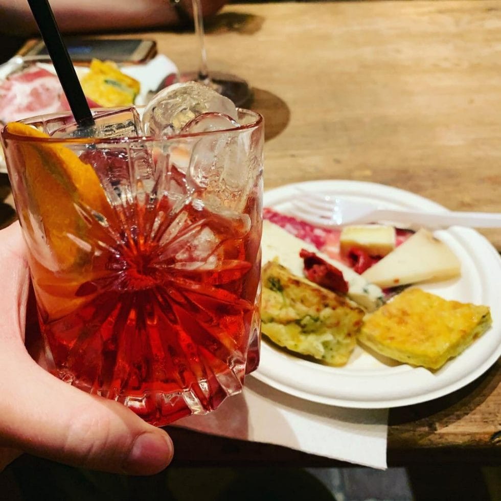 Hand holding negroni glass in front of a plate of snacks (cheese & ham etc)