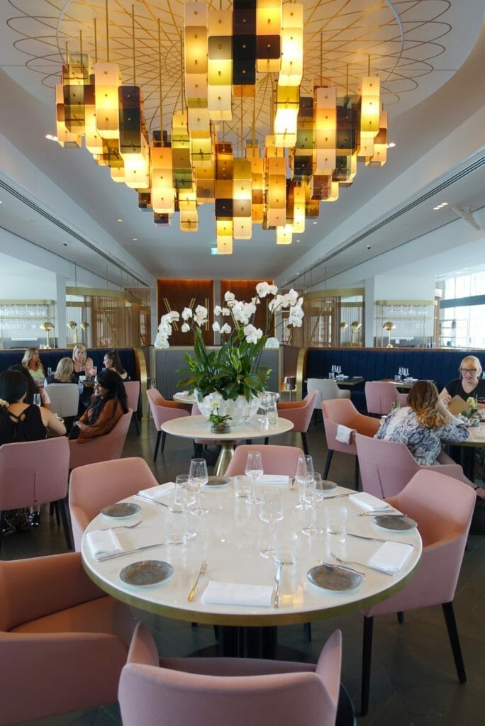 The main part of the restaurant with dusky pink chairs, round tables and a great central light fitting