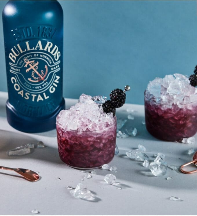 Bullards blue gin bottle in front of blue background with 2 purple bramble cocktails in front